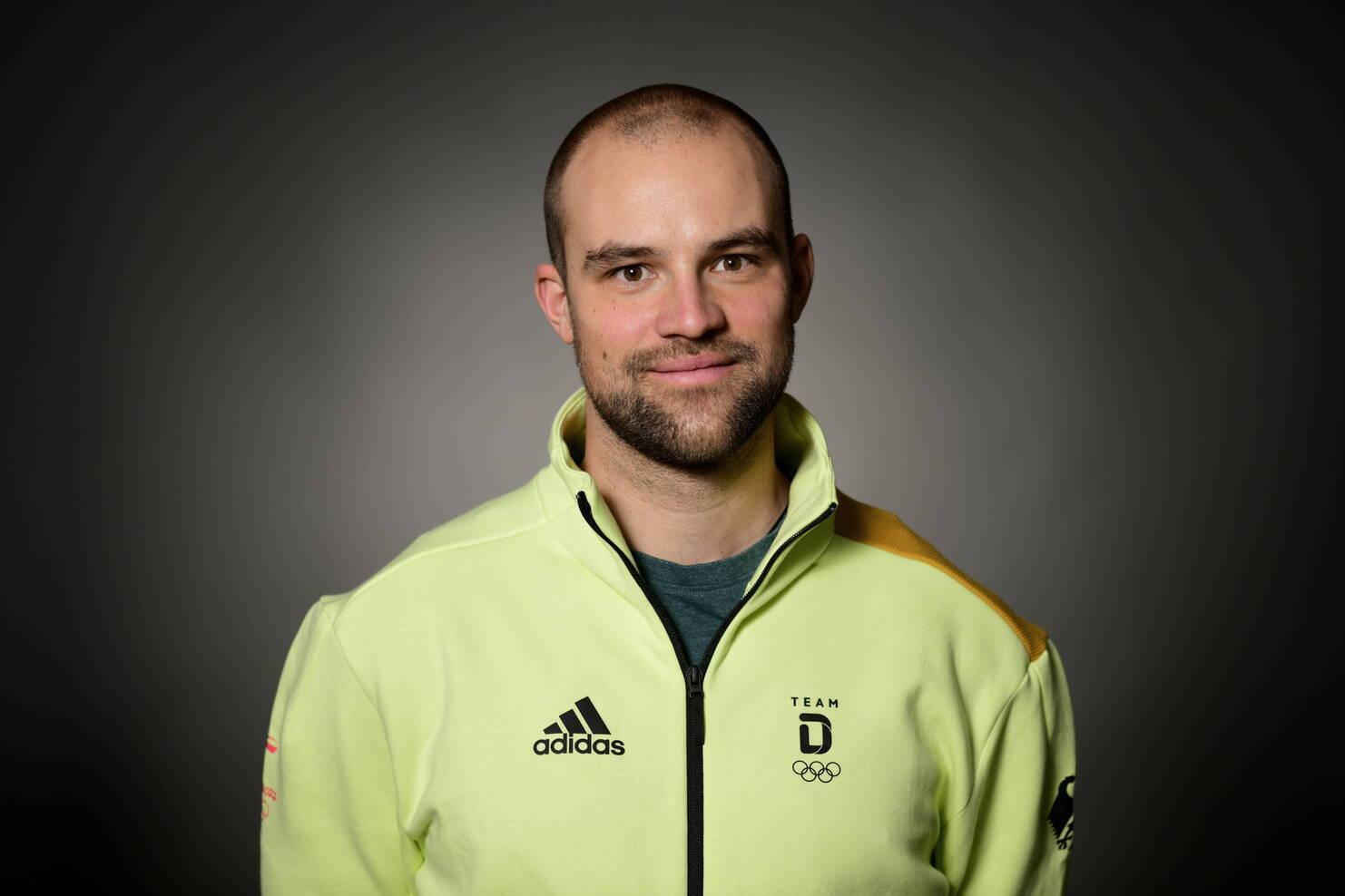 Portr&auml;t eines Mannes mit kurzen Haaren, der eine hellgr&uuml;ne Jacke mit olympischem Logo tr&auml;gt, vor einem dunklen Hintergrund.