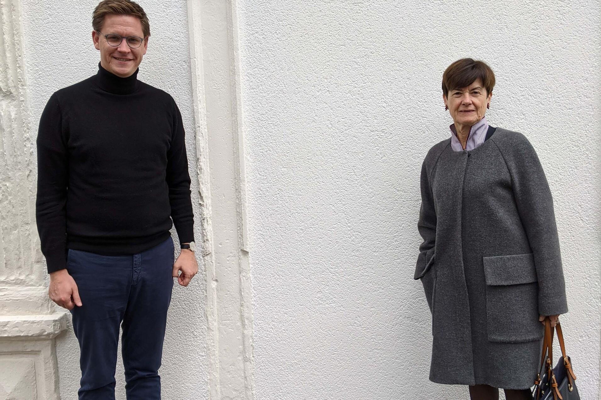 Zwei Personen stehen vor einer wei&szlig;en Wand: ein Mann in schwarzem Pullover und eine Frau in grauem Mantel.