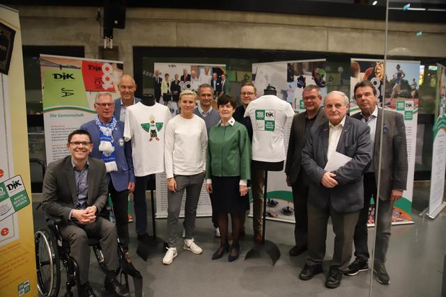 Gruppenfoto von zehn Personen, die vor einer Ausstellung mit Sportdress und Informationsplakaten stehen.