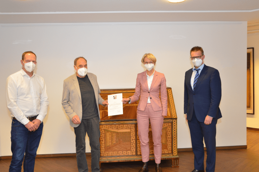 Vier Personen in Masken halten ein Dokument vor einem Kunstwerk in einem modernen Raum.