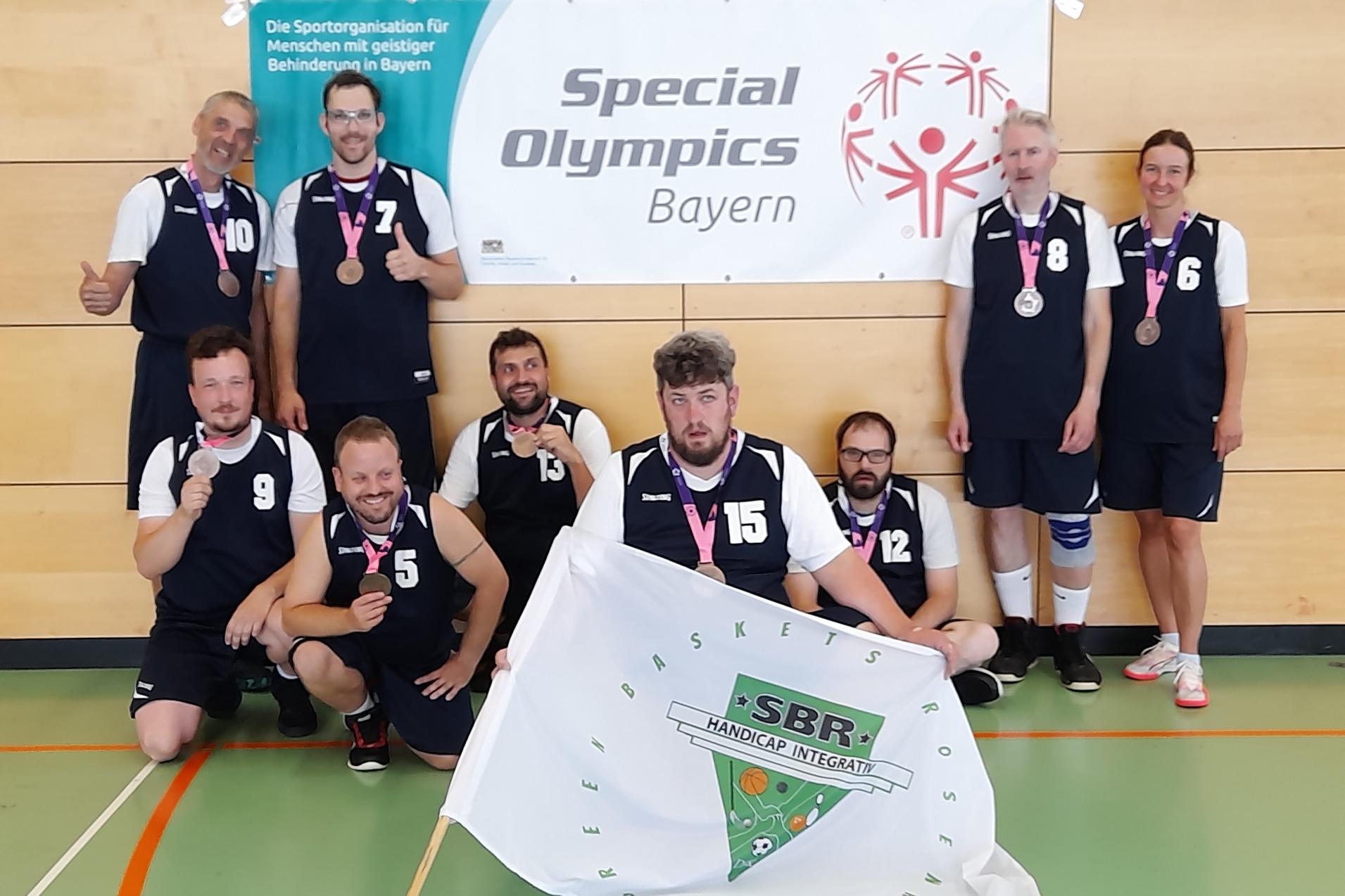 Gruppe von acht Sportlern in Basketballtrikots mit Medaillen vor Banner der Special Olympics Bayern und Fahne des Vereins.