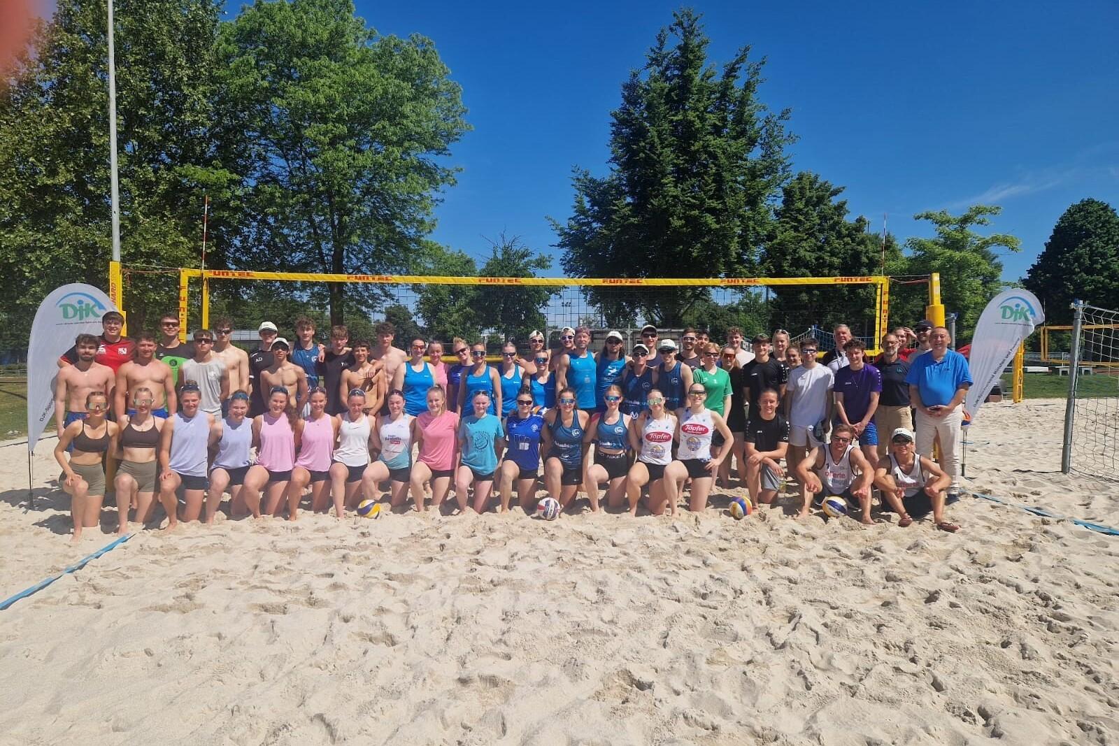 Gruppenshot von 50 Beachvolleyballspielern auf einem Sandplatz, umrahmt von B&auml;umen und einem blauen Himmel.