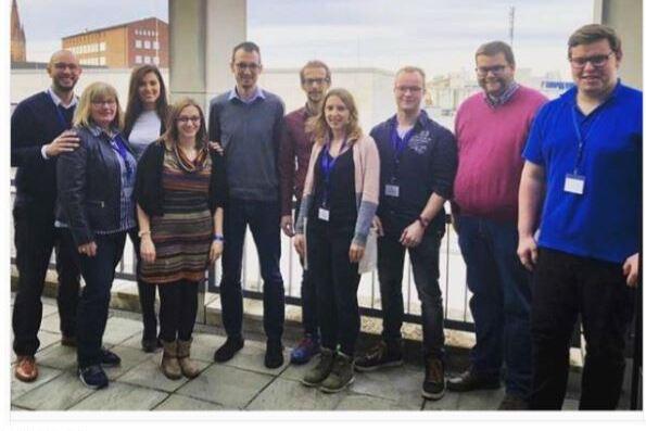 Gruppe von zehn Personen steht auf einer Terrasse mit Blick auf eine Stadtlandschaft, vor Wolken am Himmel.