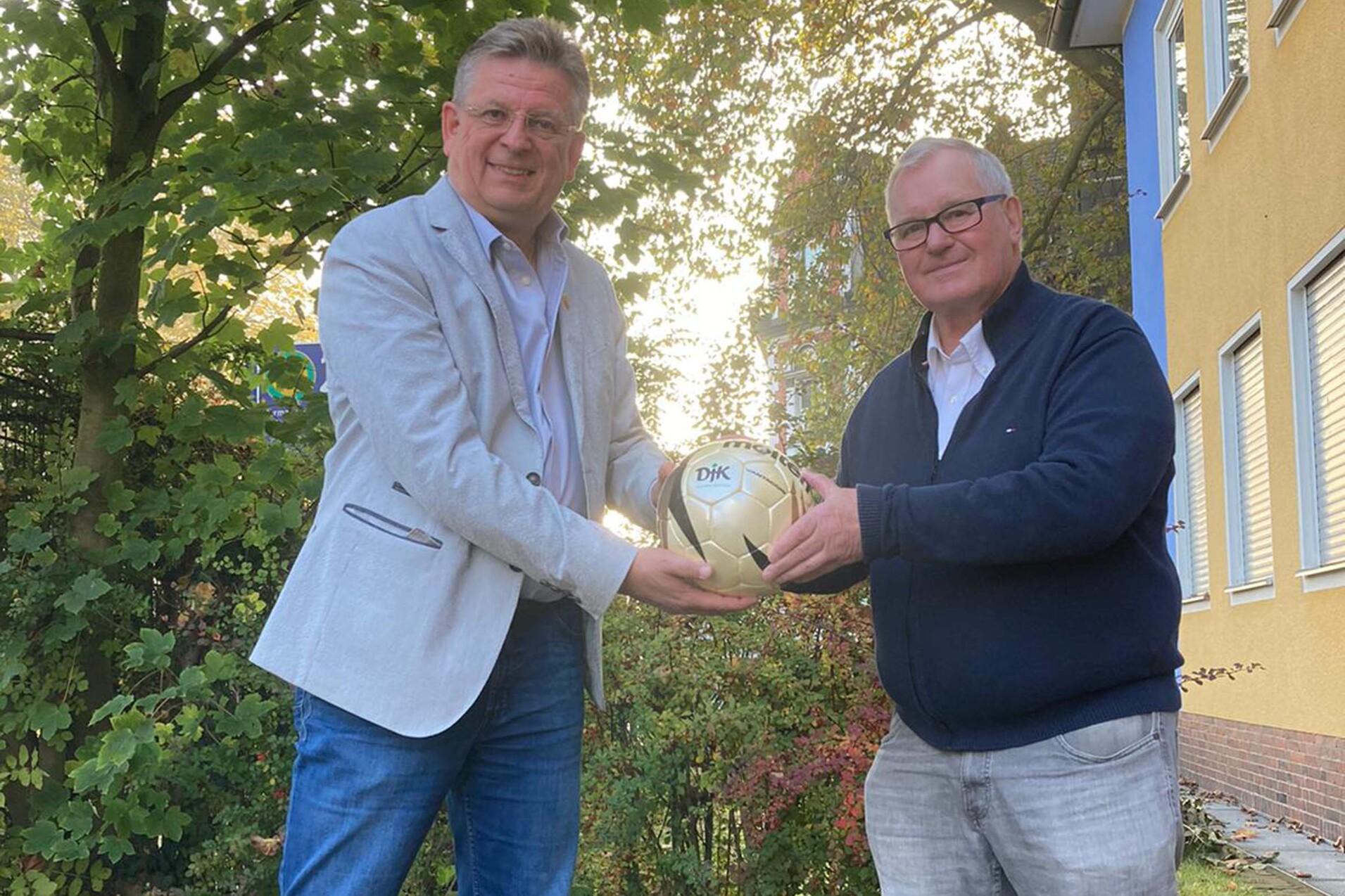 Zwei M&auml;nner &uuml;berreichen sich einen Fu&szlig;ball vor einer gr&uuml;nen Umgebung und einem gelben Geb&auml;ude im Hintergrund.