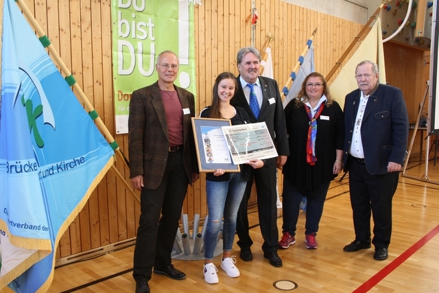 F&uuml;nf Personen stehen in einer Sporthalle, eine junge Frau h&auml;lt eine Urkunde. Im Hintergrund sind Flaggen und ein Banner sichtbar.