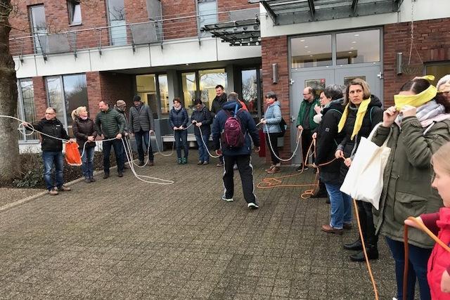 Gruppenevent im Freien: Teilnehmer halten Seile und stehen in einer Reihe vor einem Geb&auml;ude, einige tragen Augenbinden.
