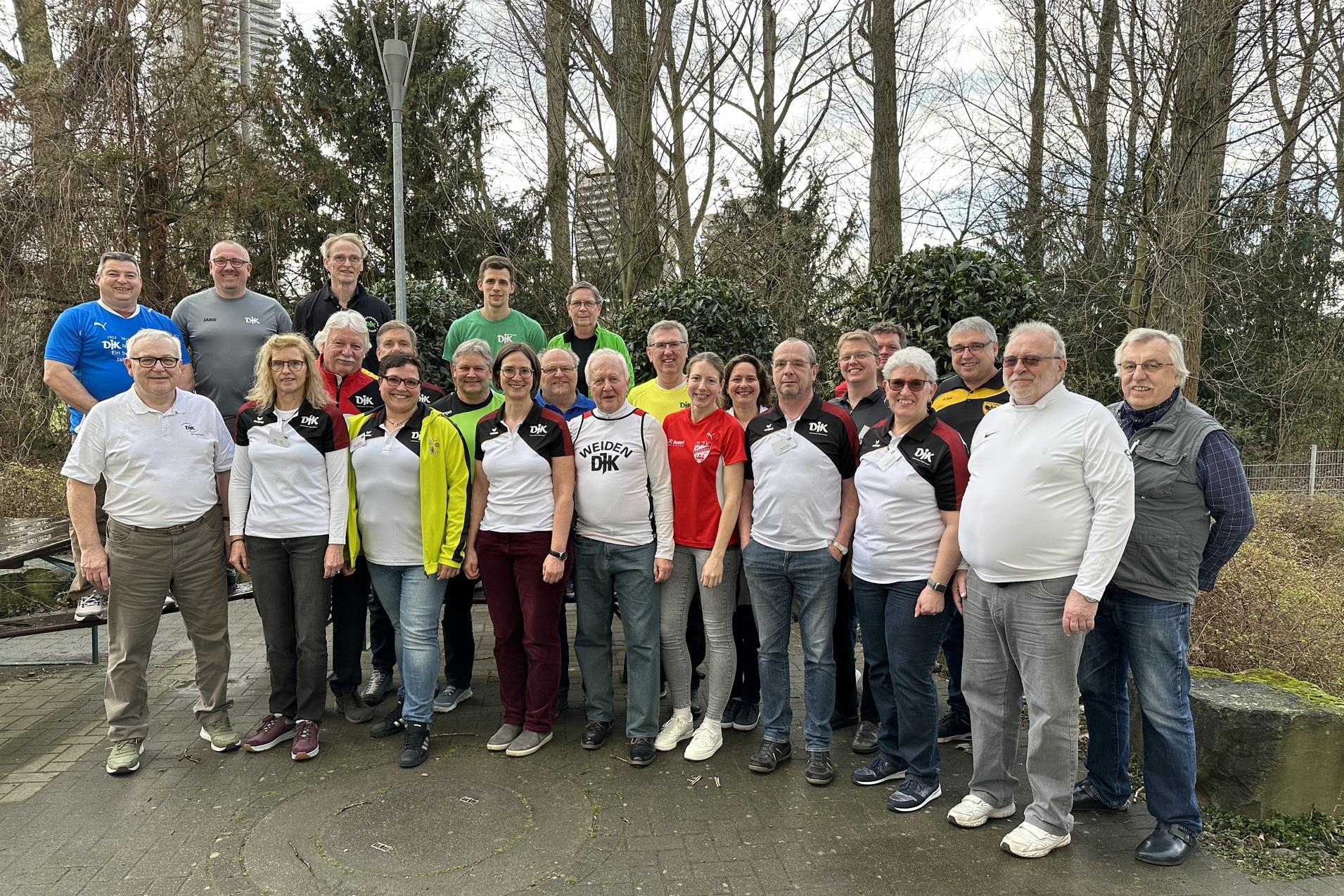 Gruppenfoto von 25 Personen in Sportkleidung, stehend in einem Park mit B&auml;umen und Laternen im Hintergrund.