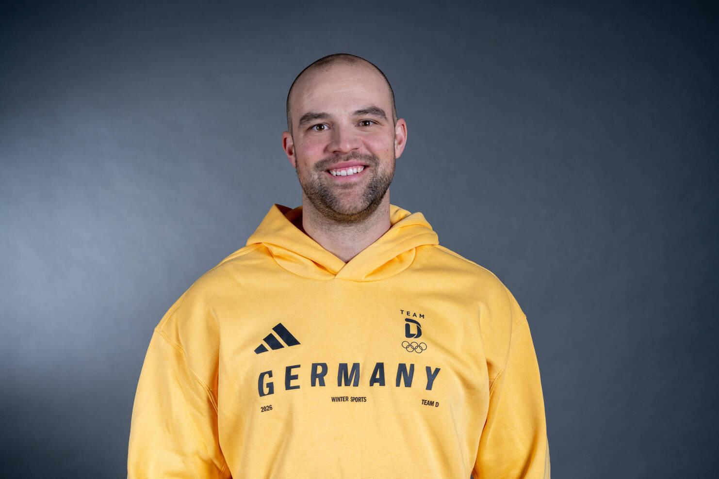 L&auml;chelnder Mann in gelbem Olympiatrainingsanzug mit Aufschrift &bdquo;Germany&ldquo; vor grauem Hintergrund.