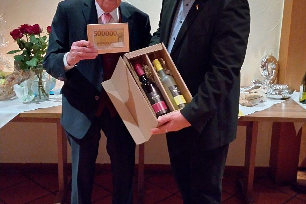 Zwei M&auml;nner stehen nebeneinander, einer h&auml;lt einen gro&szlig;en Scheck, der andere eine Geschenkverpackung mit Weinflaschen.