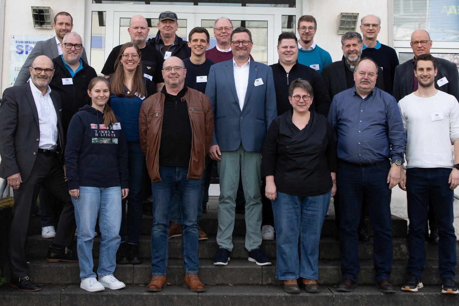Gruppenfoto von 24 Personen vor einem Geb&auml;ude, alle l&auml;cheln und tragen Namensschilder.