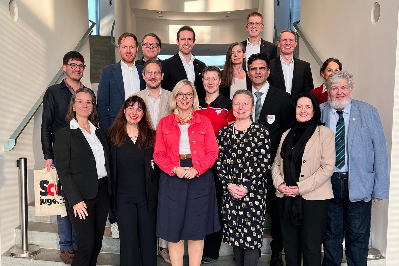 Gruppe von 15 Personen in formeller Kleidung auf einer Treppe, l&auml;chelnd und in lockerer Pose.