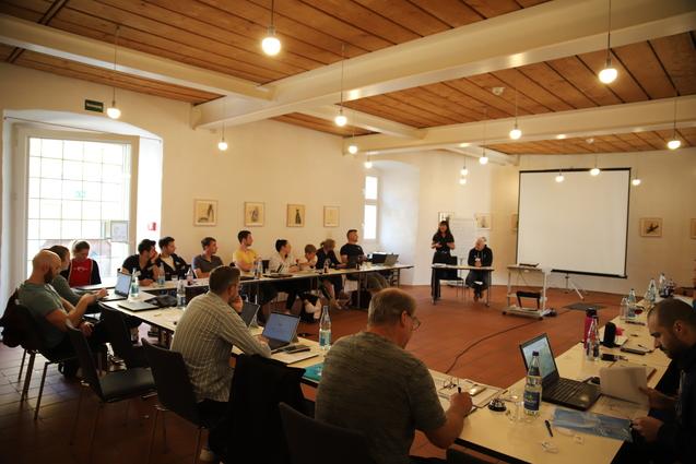 Gruppenarbeit in einem Seminarraum mit Teilnehmern an Tischen, Pr&auml;sentation an der Wand und Moderatorin vorne.