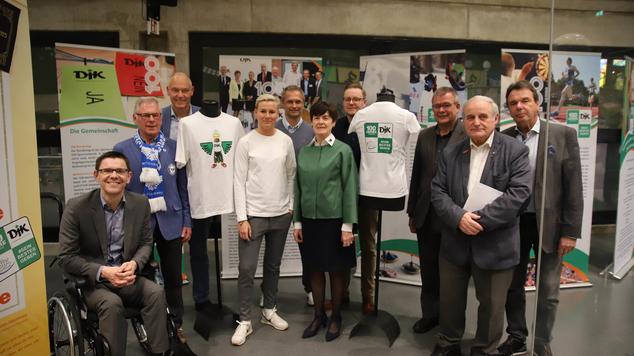 Gruppenfoto von zehn Personen, die vor einer Ausstellung mit Sportdress und Informationsplakaten stehen.