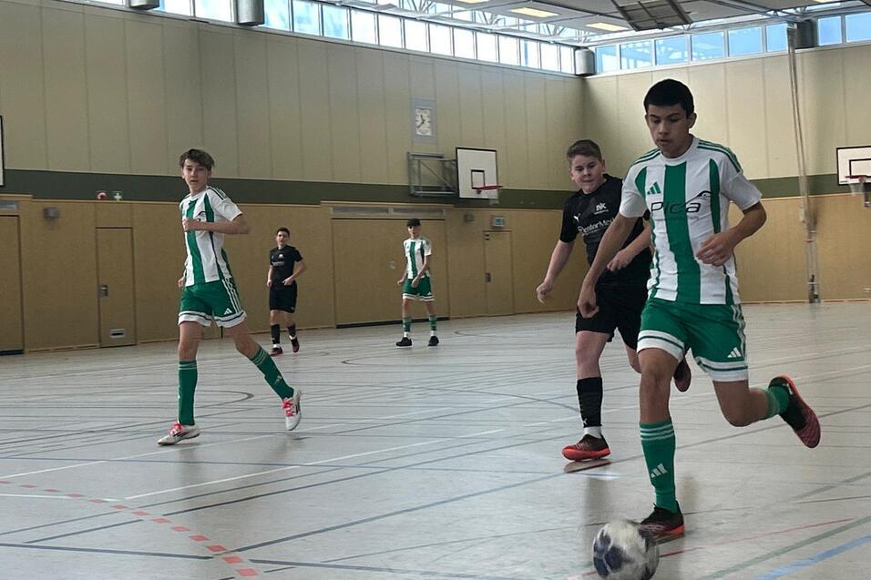 Drei Jugendliche spielen Fu&szlig;ball in einer Sporthalle, einer f&uuml;hrt den Ball, w&auml;hrend die anderen verfolgen.