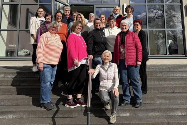 Gruppenfoto von 18 Frauen, die auf Treppen vor einem gro&szlig;en Fenster stehen, mit unterschiedlichen Kleidungsstilen und freundlichen Gesichtsausdr&uuml;cken.