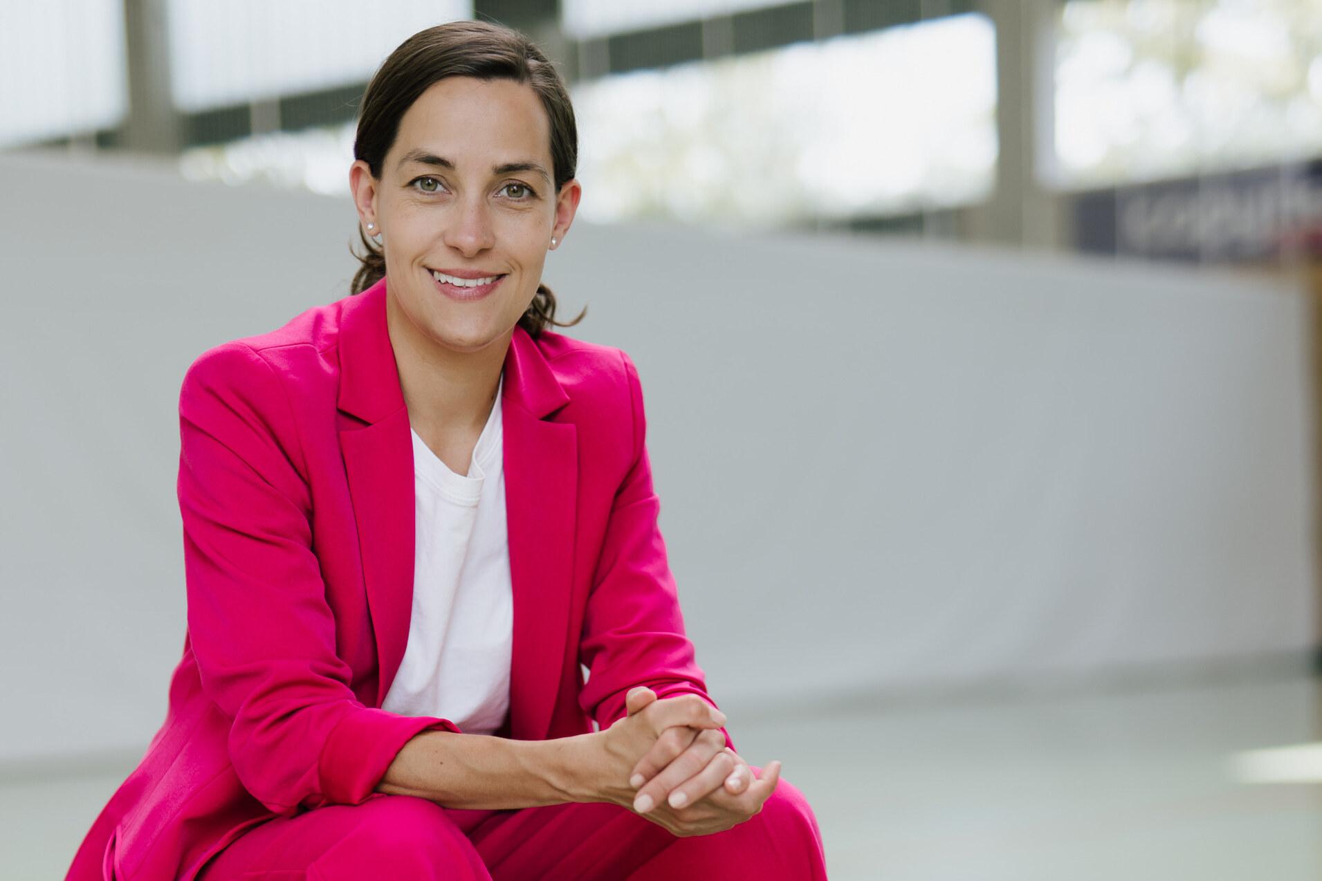 Frau in pinkem Anzug sitzt auf einer Bank, umgeben von Sportb&auml;llen: Basketball, Fu&szlig;- und Volleyball.