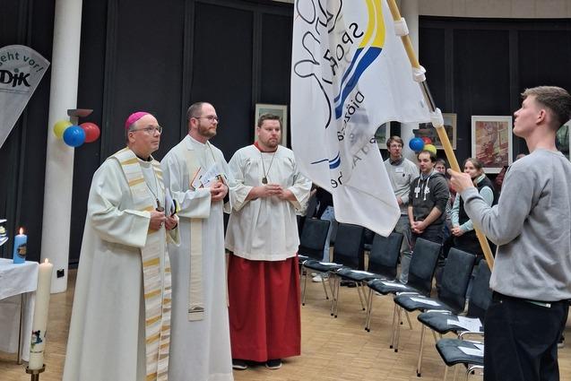 Zwei Geistliche und ein junger Mann halten eine Flagge w&auml;hrend einer Veranstaltung in einem geschlossenen Raum.