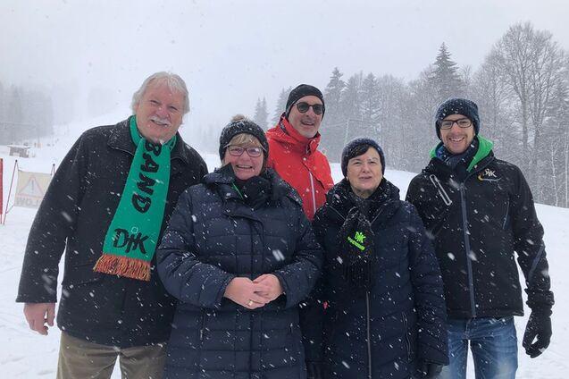 F&uuml;nf Personen stehen im Schneefall auf einer verschneiten Fl&auml;che mit B&auml;umen im Hintergrund, alle in winterlicher Kleidung.