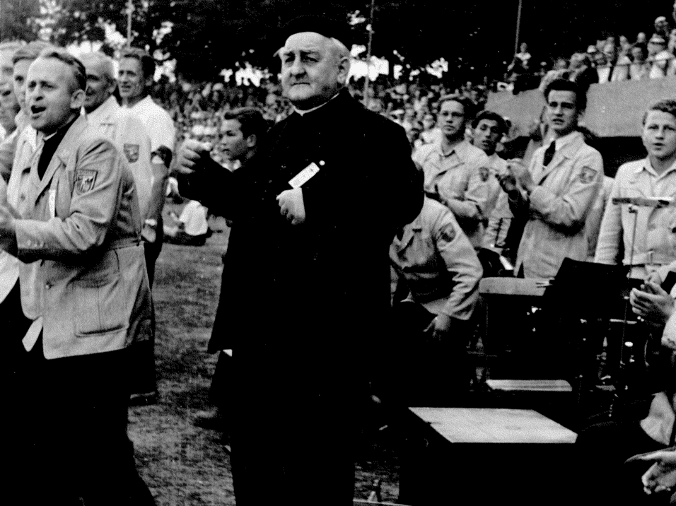 Gruppenszene auf einem Sportplatz mit applaudierenden M&auml;nnern in Uniform und einem &auml;lteren Mann im Vordergrund.
