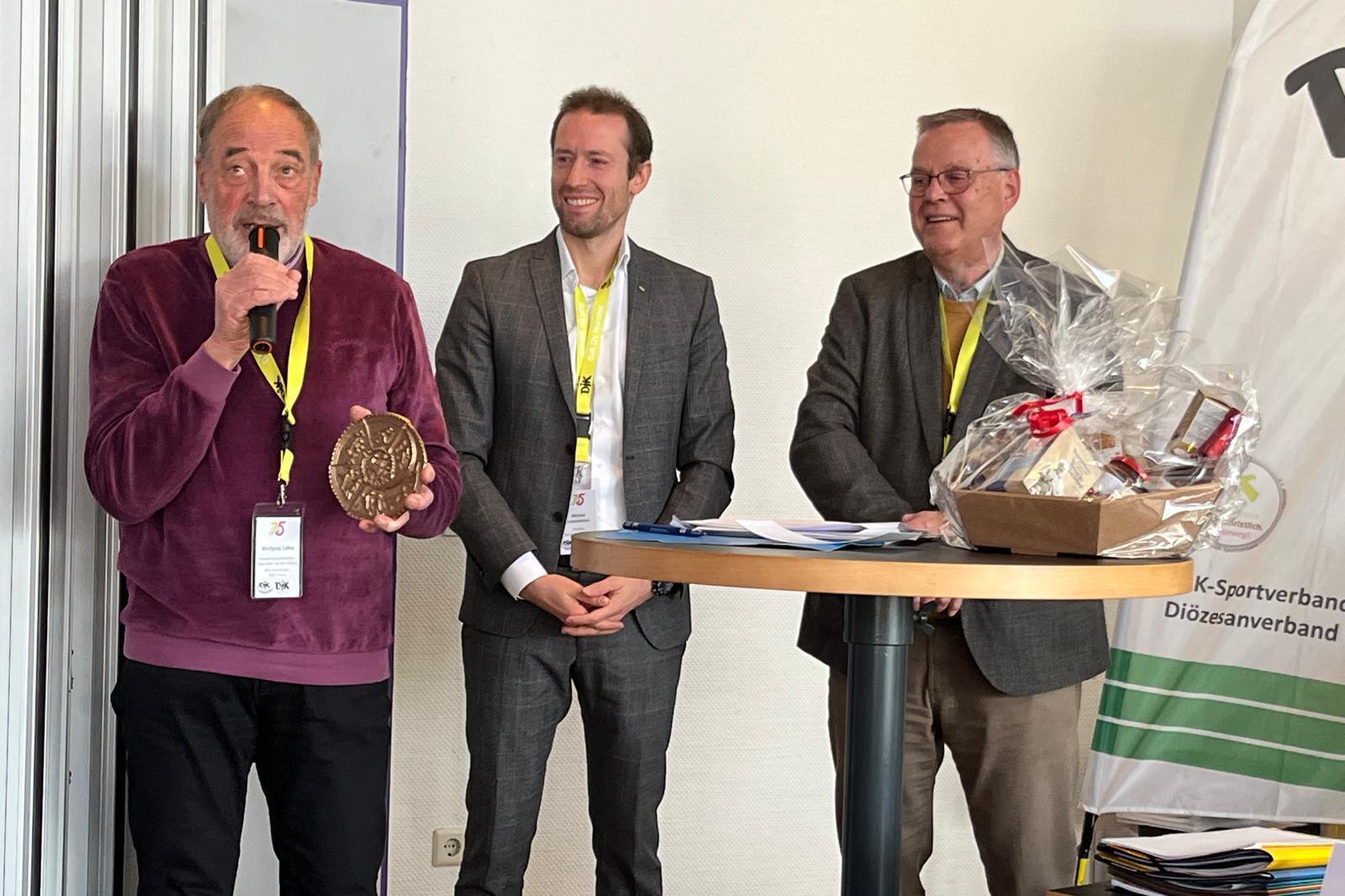 Drei M&auml;nner stehen an einem Tisch, einer h&auml;lt ein Mikrofon und eine Auszeichnung, w&auml;hrend die anderen l&auml;cheln.