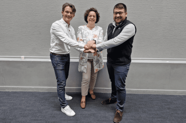 Drei Personen stehen zusammen und halten ihre H&auml;nde in der Mitte; sie l&auml;cheln und stehen vor einer grauen Wand.