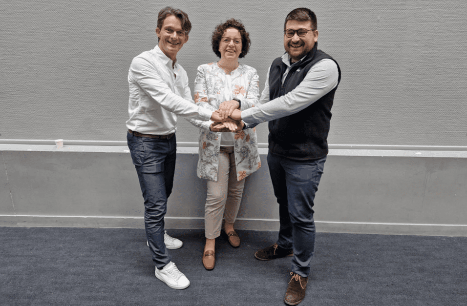 Drei Personen stehen zusammen und halten ihre H&auml;nde in der Mitte; sie l&auml;cheln und stehen vor einer grauen Wand.