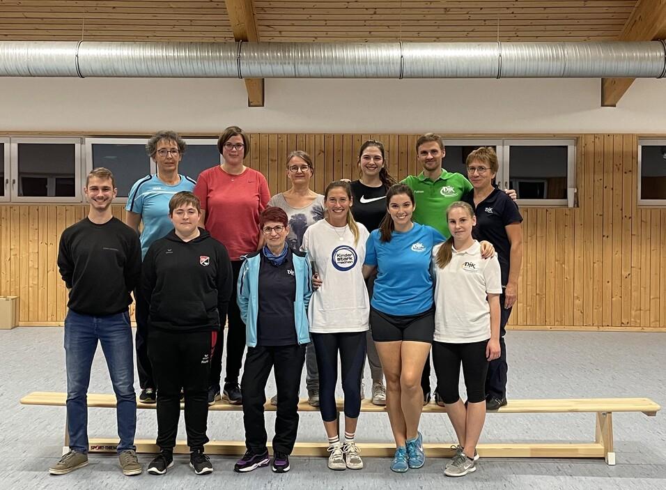 Gruppenfoto von 14 Personen in Sportkleidung, auf einer Holzbank in einem hellen Raum mit Holzvert&auml;felung.
