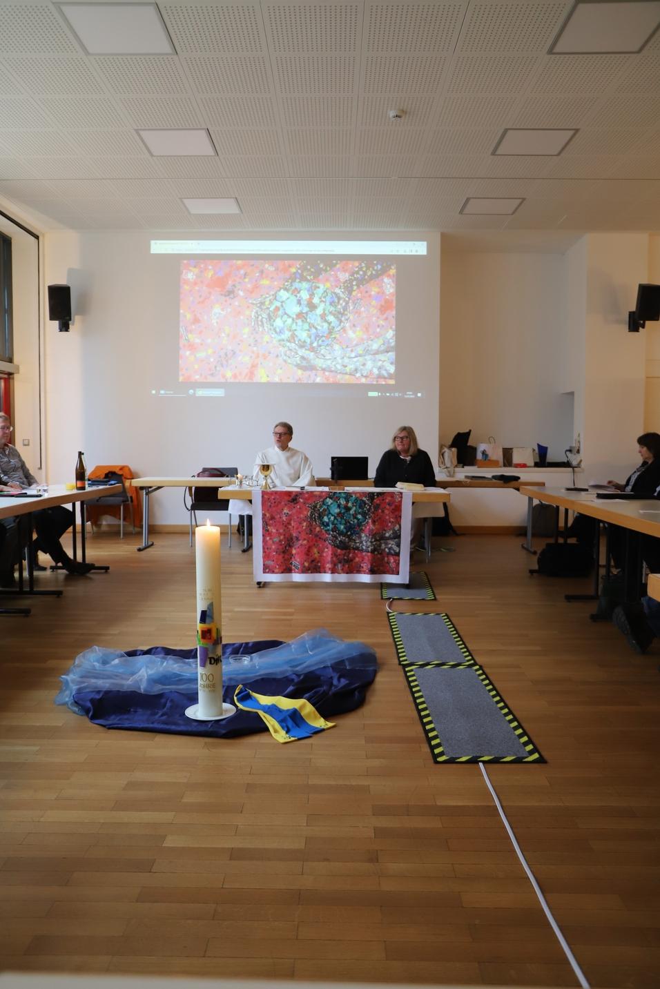 Raum mit Tischen, einer Kerze in der Mitte, und einer Pr&auml;sentation an der Wand. Teilnehmer in Diskussion.