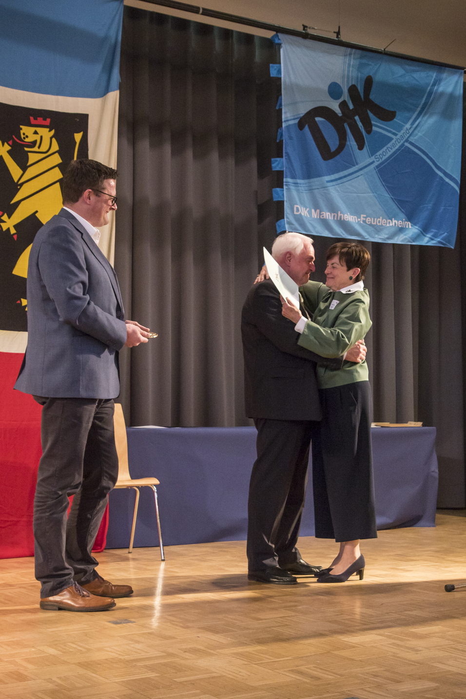 Zwei Personen umarmen sich auf einer B&uuml;hne, w&auml;hrend ein dritter Mensch ihnen zusieht. Im Hintergrund ein Banner und Fahne.