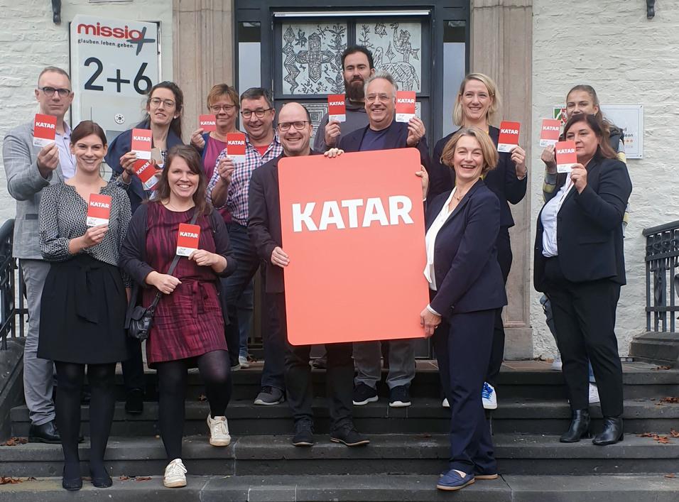 Gruppe von Personen auf Treppe, alle halten Karten mit der Aufschrift &bdquo;KATAR&ldquo; in der Hand.