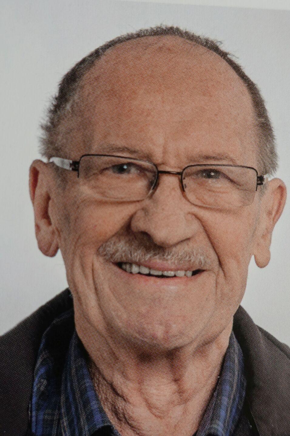 L&auml;chelnder &auml;lterer Mann mit Brille und kurz geschnittenem Haar, tr&auml;gt eine kariierte Bluse und einen dunklen Oberbekleidungsst&uuml;ck.