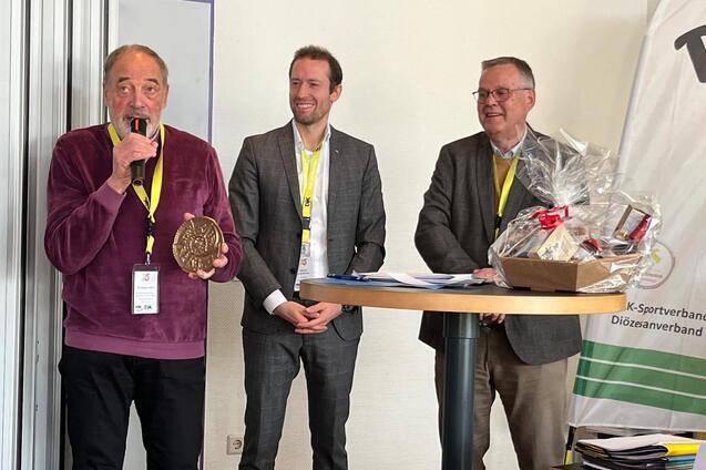 Drei M&auml;nner stehen an einem Tisch, einer h&auml;lt ein Mikrofon und eine Auszeichnung, w&auml;hrend die anderen l&auml;cheln.