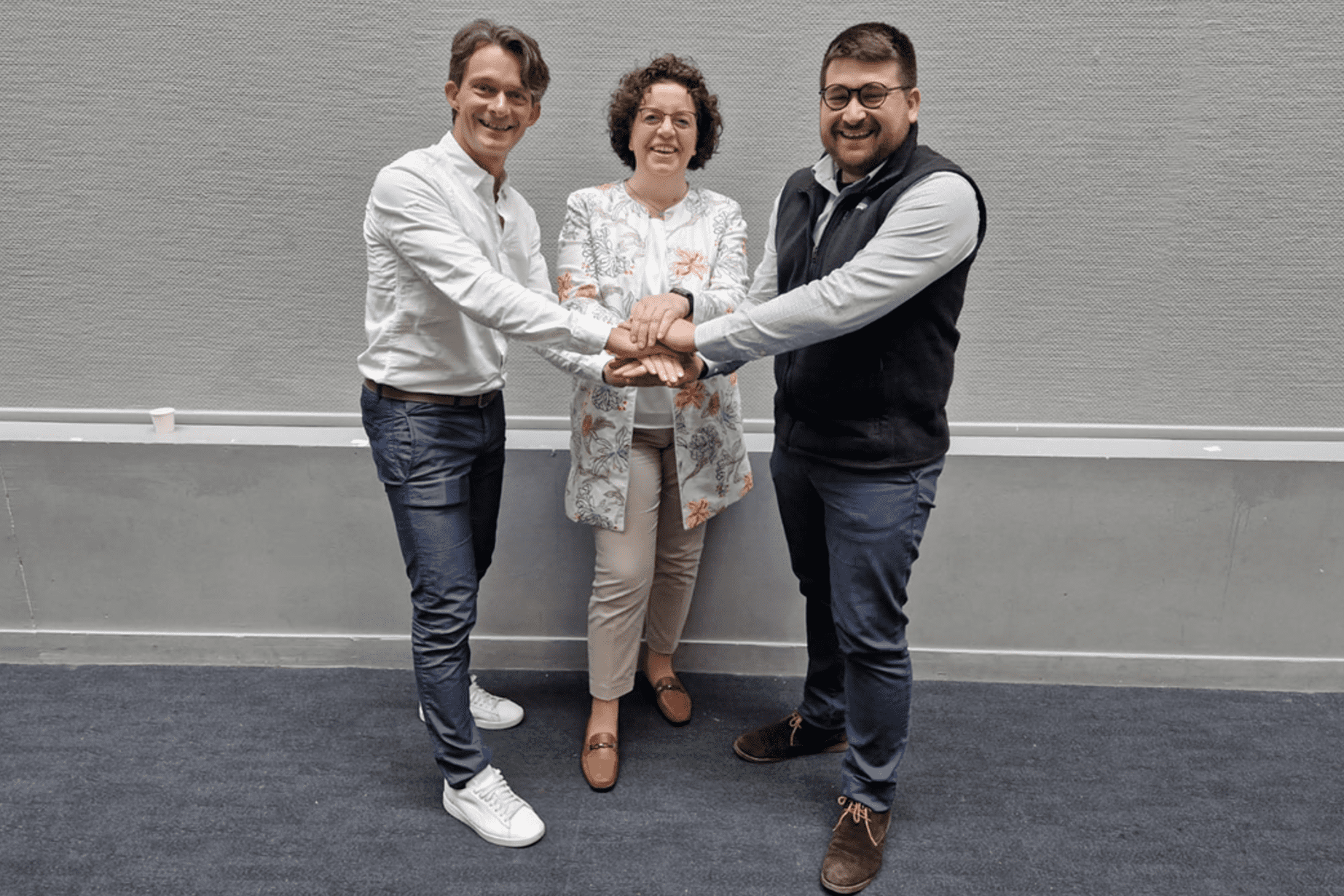 Drei Personen stehen zusammen und halten ihre H&auml;nde in der Mitte; sie l&auml;cheln und stehen vor einer grauen Wand.