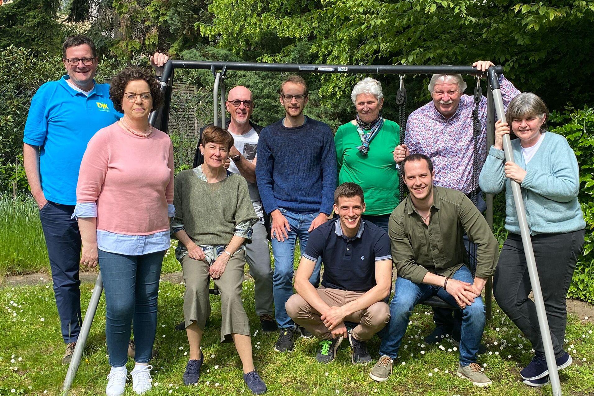 Gruppe von zehn Personen l&auml;chelnd im Freien, stehend und sitzend vor einer Schaukellage in einem Garten.
