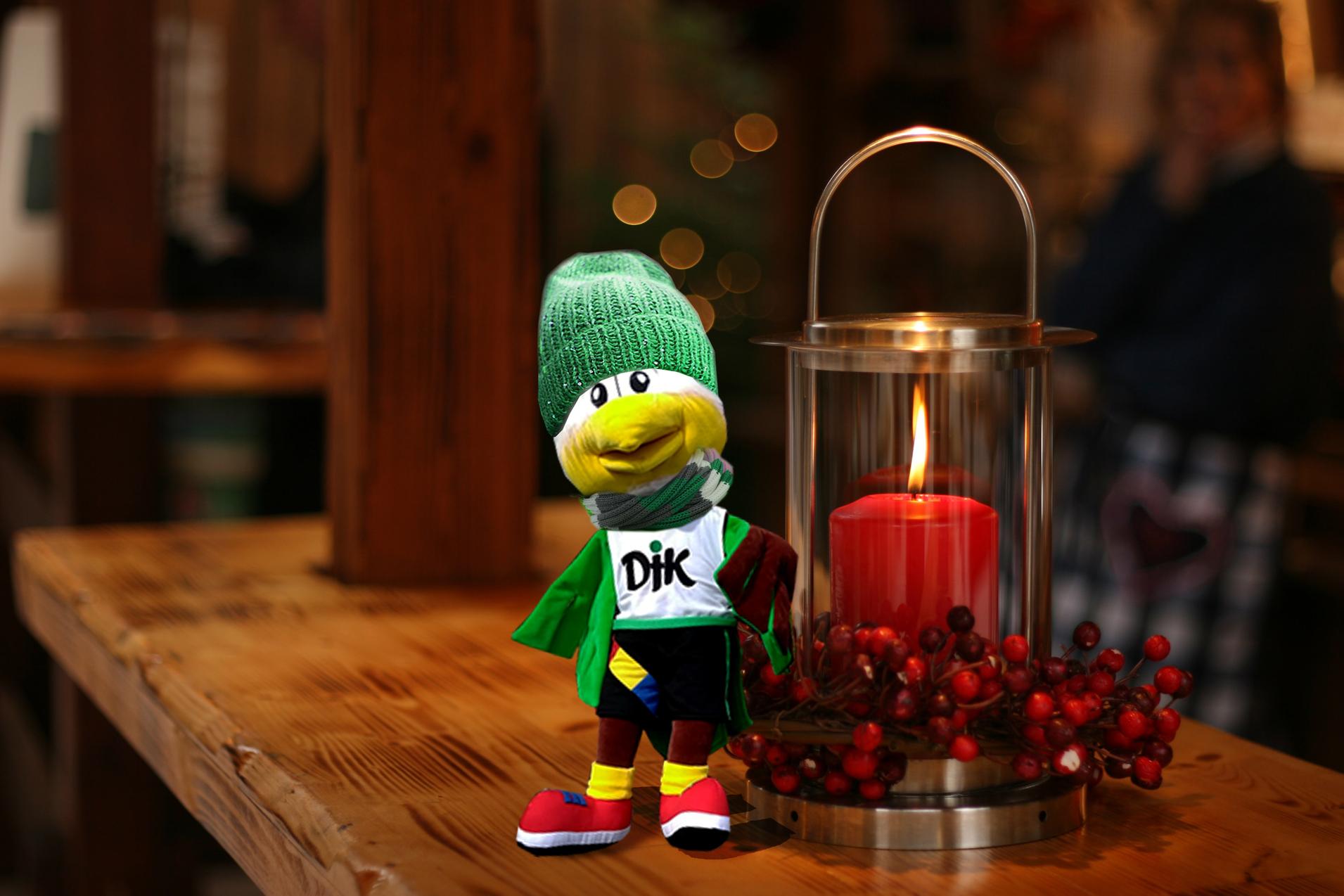 Pl&uuml;schfigur mit gr&uuml;nem M&uuml;tze steht vor einer Laterne mit rot leuchtender Kerze und Dekoration aus roten Beeren.