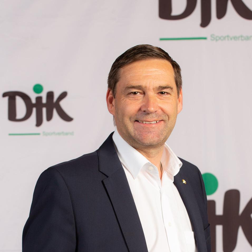 Mann in Anzug h&auml;lt einen bunten Fu&szlig;ball vor einem Hintergrund mit dem Logo des DJK Sportverbands.
