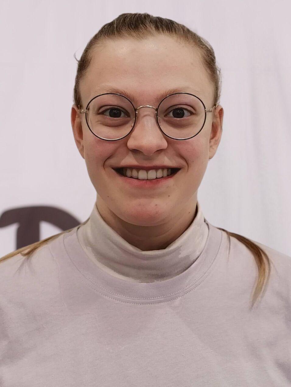 L&auml;chelnde Person mit Brille, tr&auml;gt ein graues Oberteil, vor einem wei&szlig;en Hintergrund mit DJK-Logo.