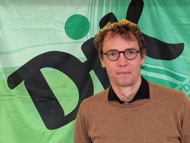 Mann mit Brille steht vor einem gr&uuml;nen Hintergrund mit dem Wort "Dix" in schwarzer Schrift.