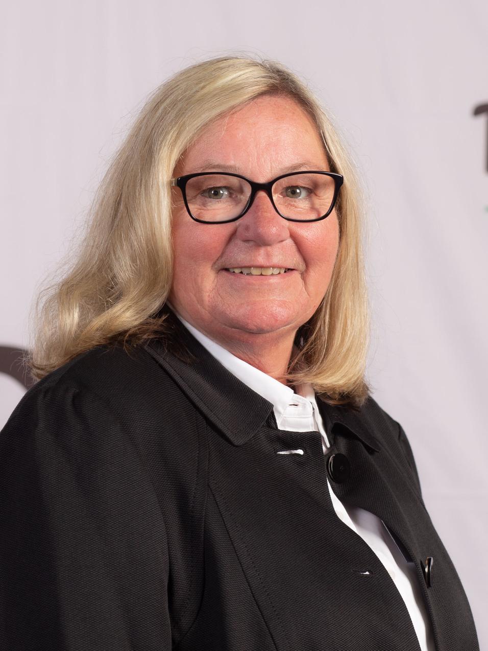 Portrait einer l&auml;chelnden Frau mit blonden Haaren und Brille vor einem DJK-Hintergrund.