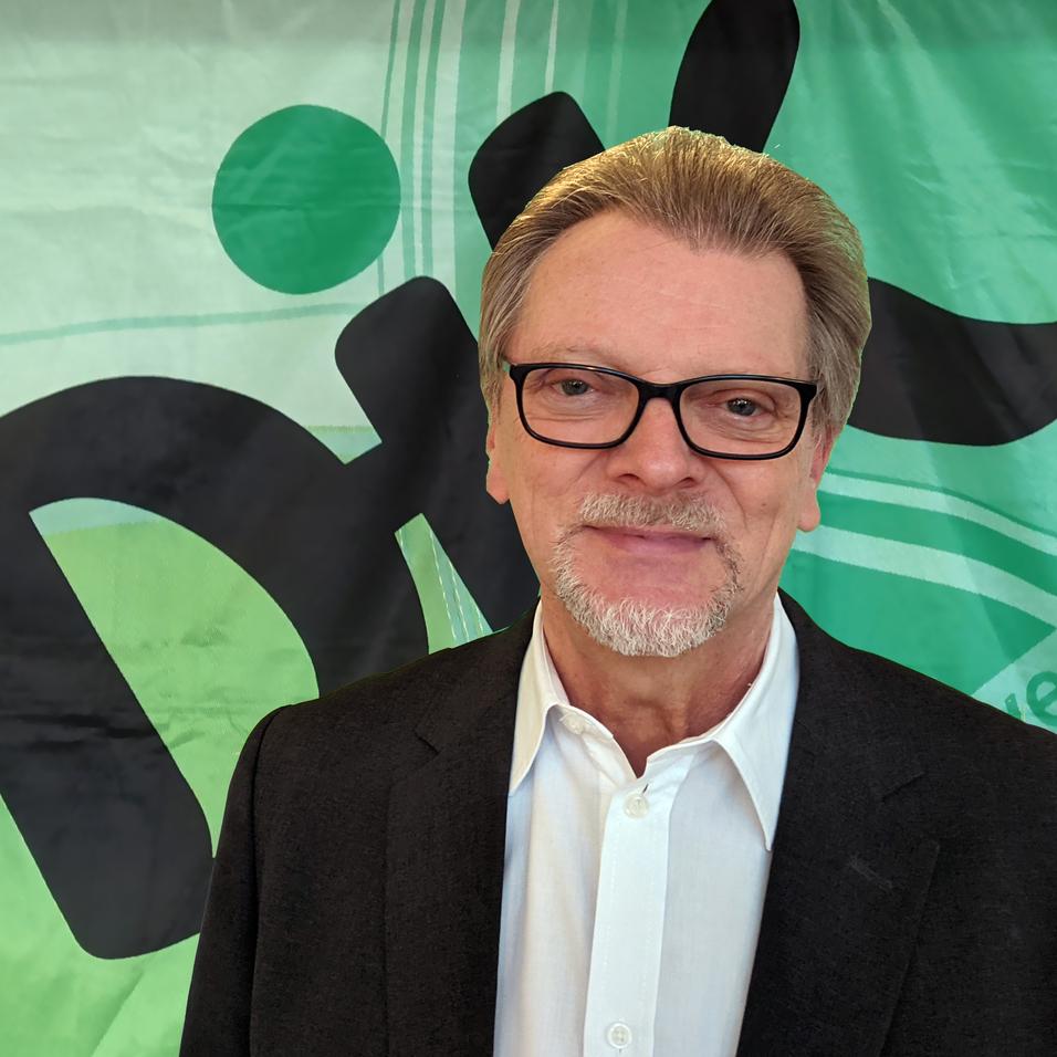 Portr&auml;t eines Mannes mit Brille in Anzug vor einem gr&uuml;nen Banner mit schwarzen und blauen grafischen Elementen.