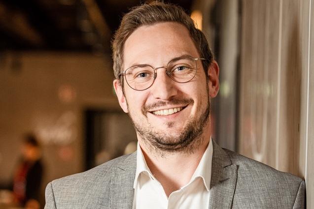 L&auml;chelnder Mann mit Brille und grauem Anzug, steht an einer Wand in einer modernen, hellen Umgebung.