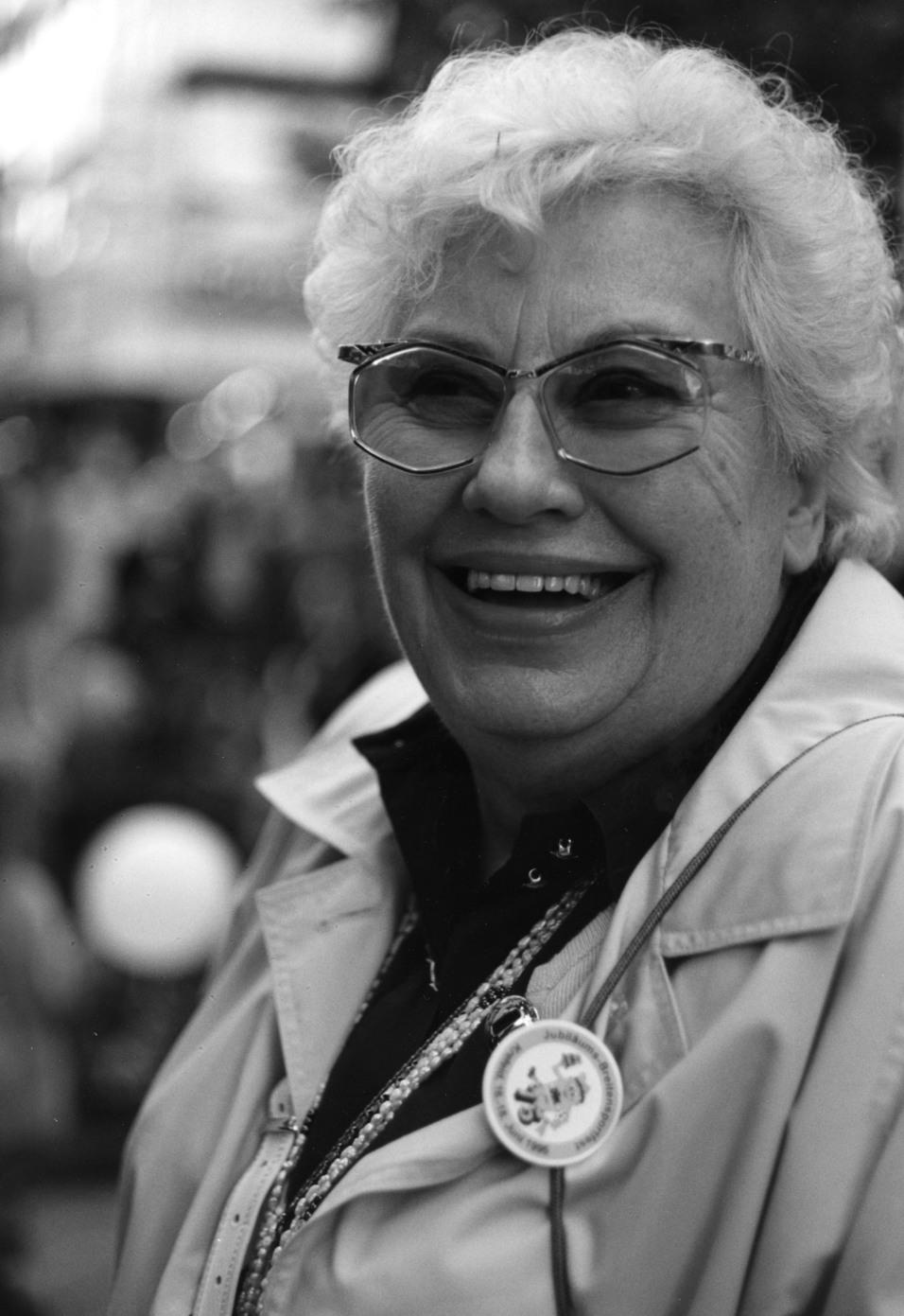 L&auml;chelnde &auml;ltere Frau mit Brille und einem Abzeichen, aufgenommen in einer lebhaften Umgebung.