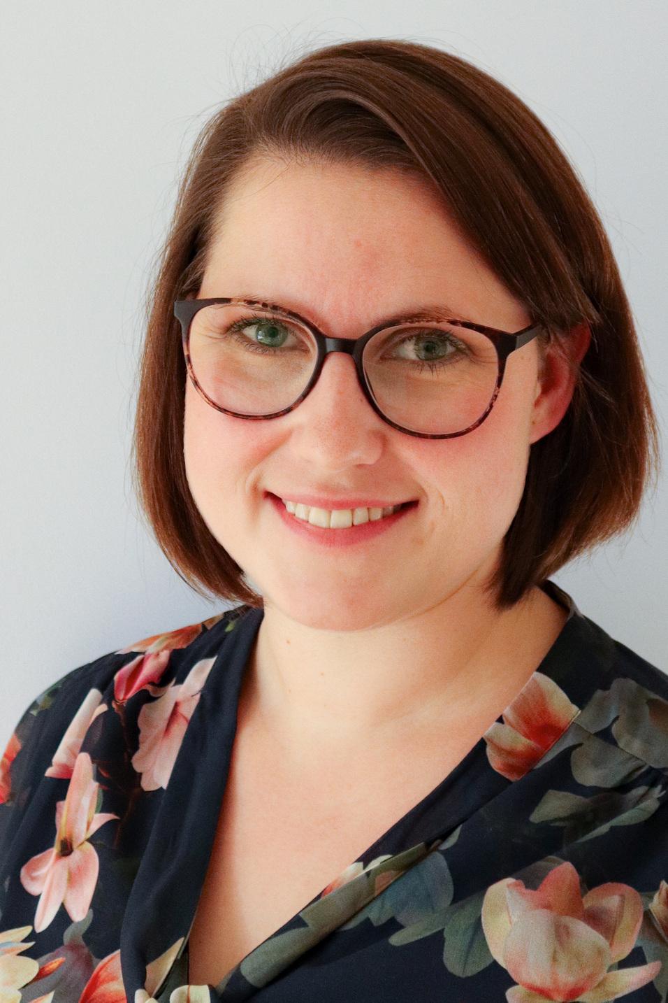 Portr&auml;t einer Frau mit kurzen, braunen Haaren und Brille, die ein blumiges Oberteil tr&auml;gt, vor neutralem Hintergrund.