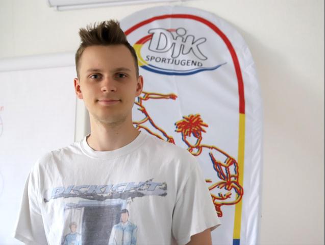 Junger Mann in einem wei&szlig;en T-Shirt vor einem Banner der DJK Sportjugend mit sportlichen Motiven.