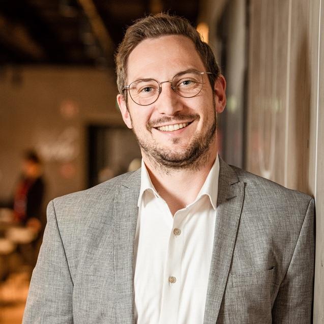 L&auml;chelnder Mann mit Brille und grauem Anzug, steht an einer Wand in einer modernen, hellen Umgebung.