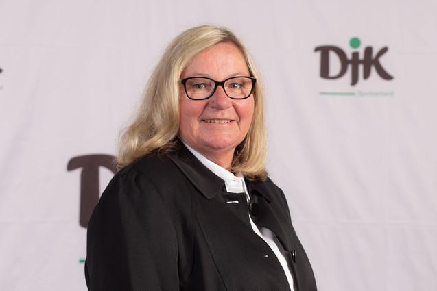 Portrait einer l&auml;chelnden Frau mit blonden Haaren und Brille vor einem DJK-Hintergrund.