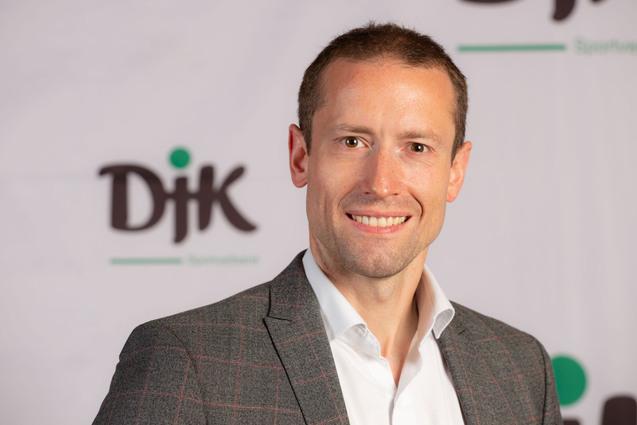 Portr&auml;t eines Mannes in kariertem Blazer vor einem DJK-Logo auf einem hellen Hintergrund. Er l&auml;chelt freundlich.