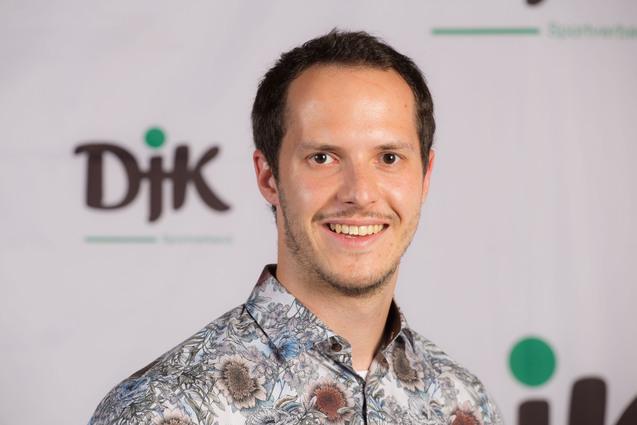 L&auml;chelnder Mann mit kurzen Haaren in einem blumigen Hemd vor einem DJK-Logo auf einer neutralen Hintergrundwand.