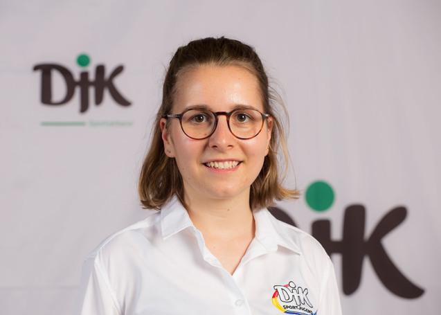 Portr&auml;t einer jungen Frau mit Brille, l&auml;chelnd, in einem wei&szlig;en Hemd vor einer DJK-Logowand.