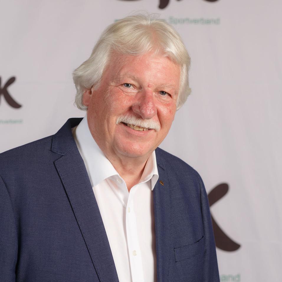 Portr&auml;t eines Mannes mit grauen Haaren und Schnurrbart, der in einem Sakko vor einem DIk-Logo steht.